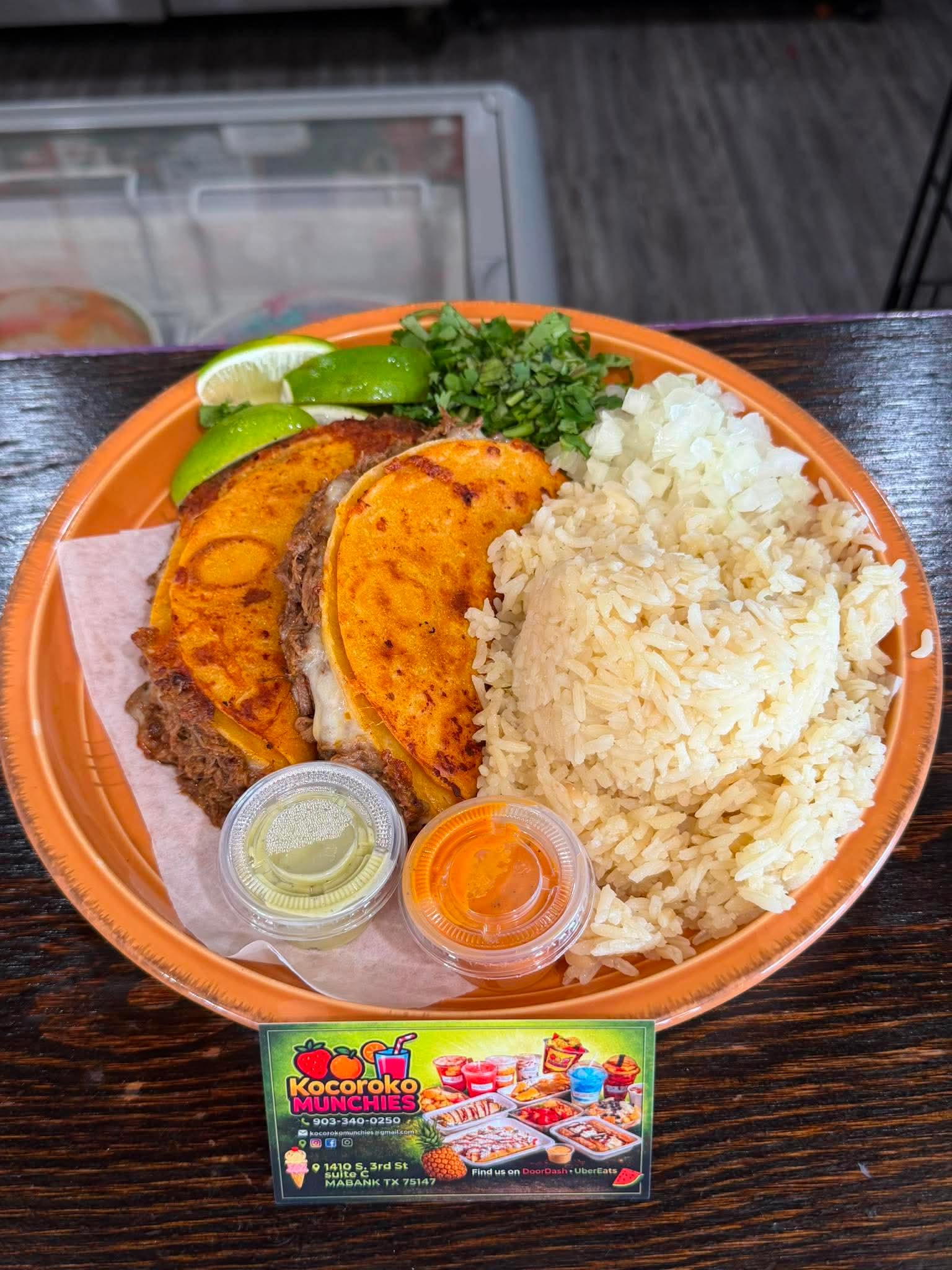 Plate with two tacos, rice, lime, cilantro, and sauces on an orange dish, on a dark wooden table.