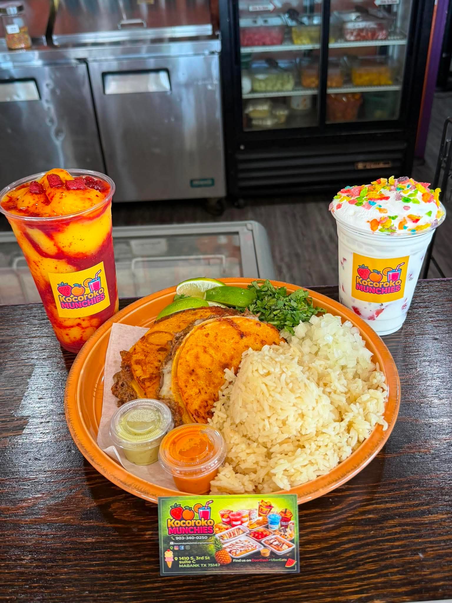 Plate with rice, fried chicken, lime, and broccoli, two colorful drinks with fruit toppings, and a business card on a dark wooden table.