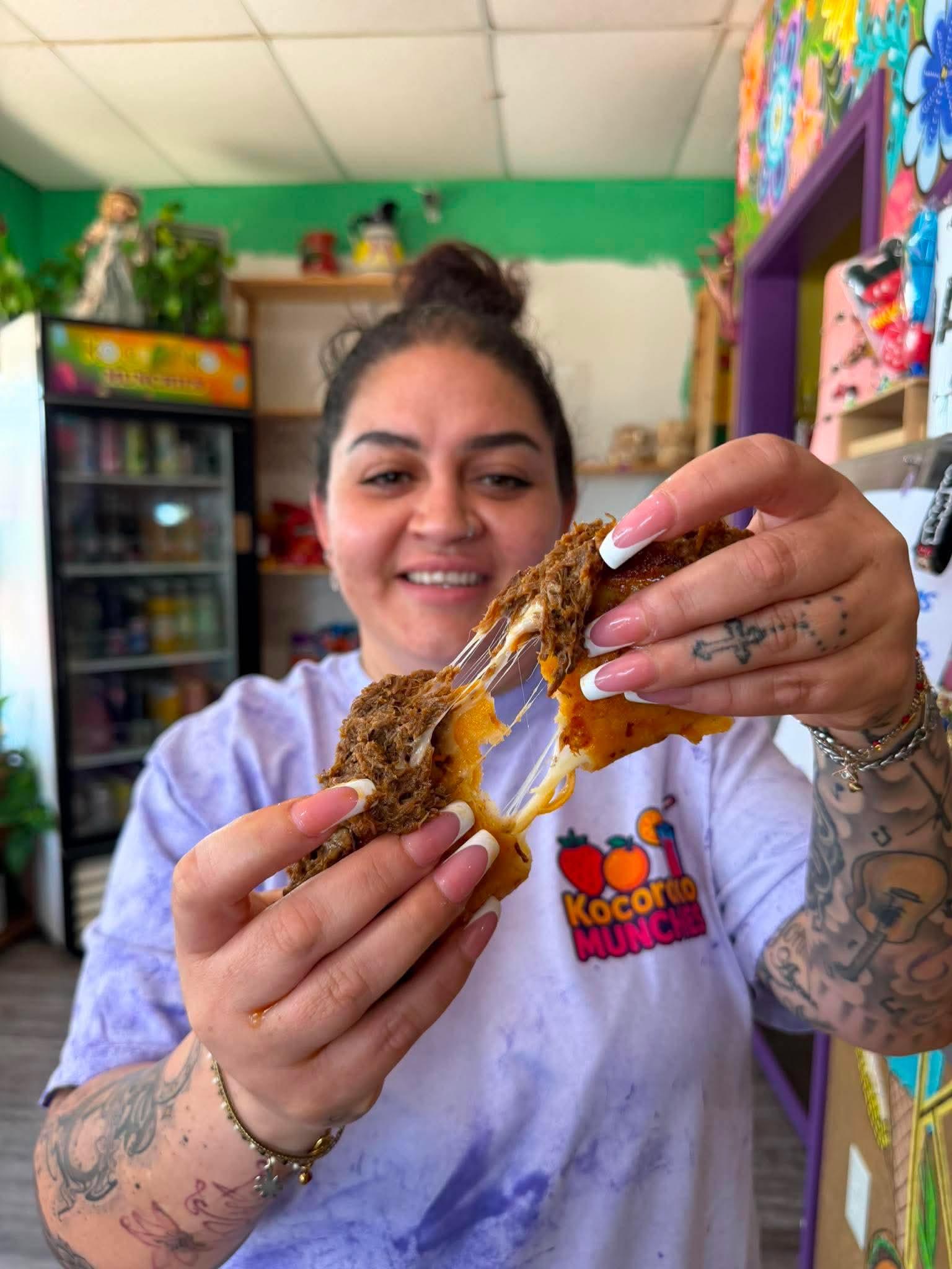 Young woman with tattoos holding a fried snack with cheese, smiling in a colorful store, wearing a white shirt with a logo.
