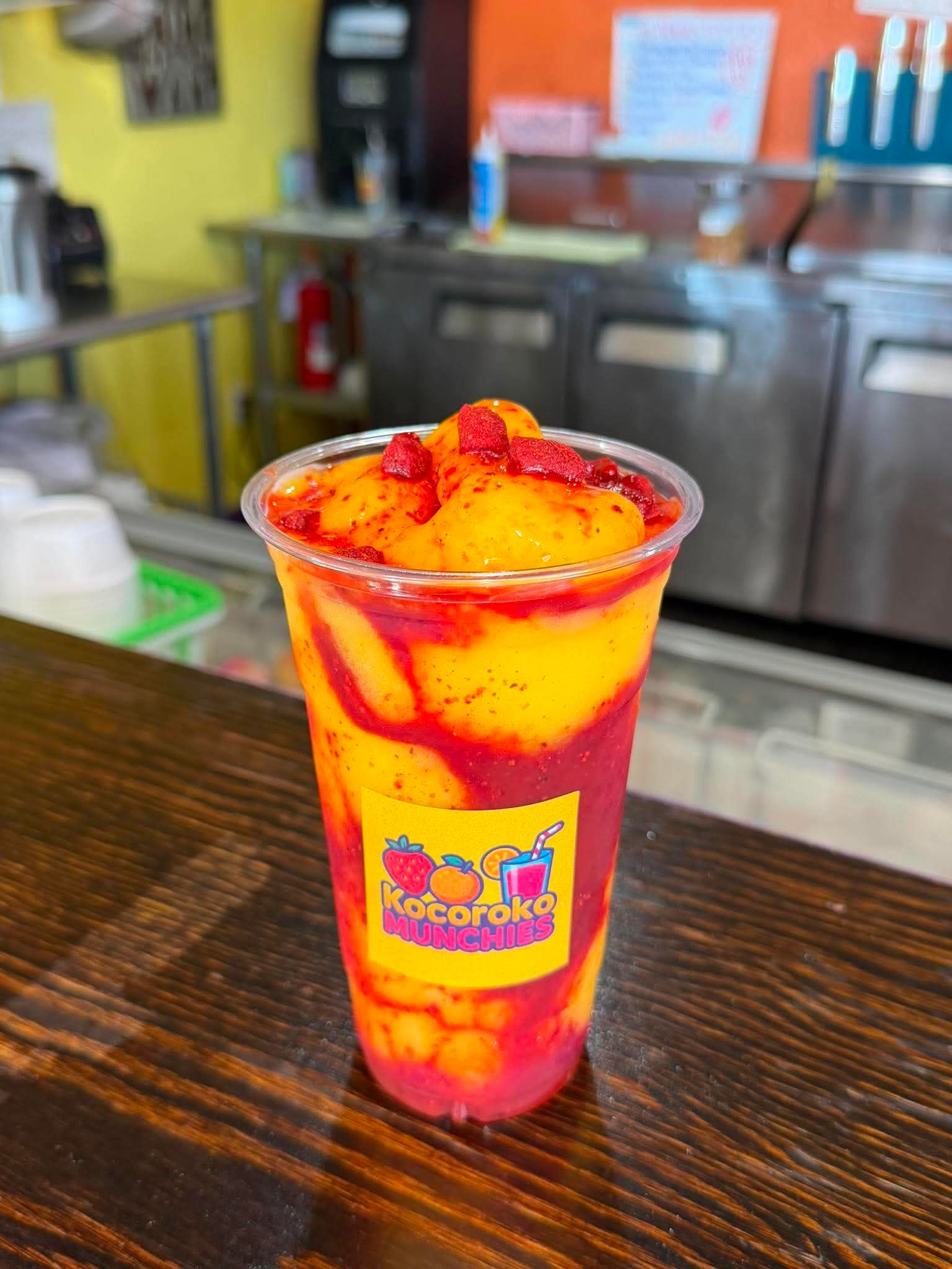 A colorful fruit drink with mango and berry pieces in a clear cup on a wooden table, with a kitchen background.