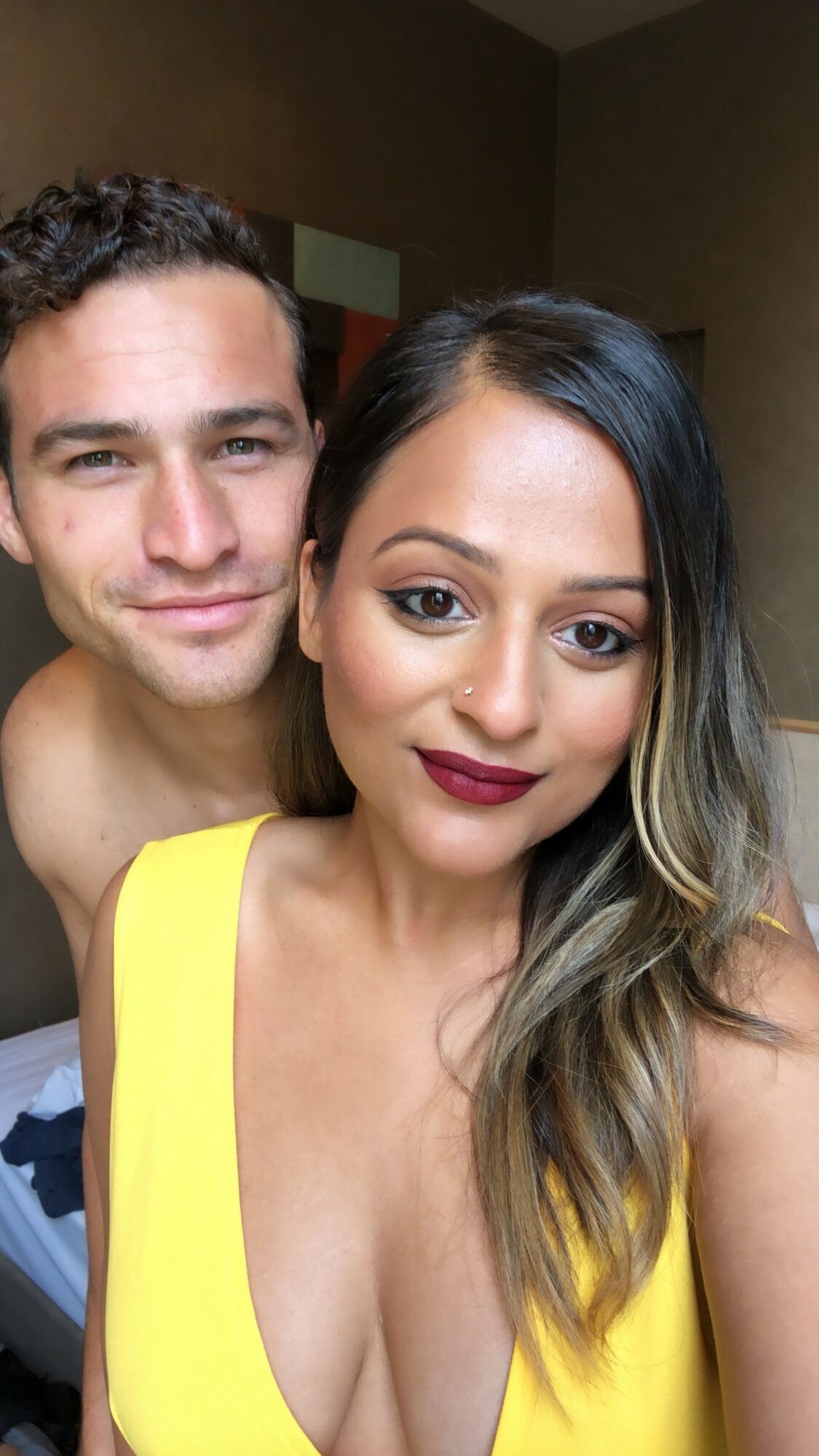 Two people, a man and a woman, pose closely indoors, woman wearing a yellow top, both smiling.