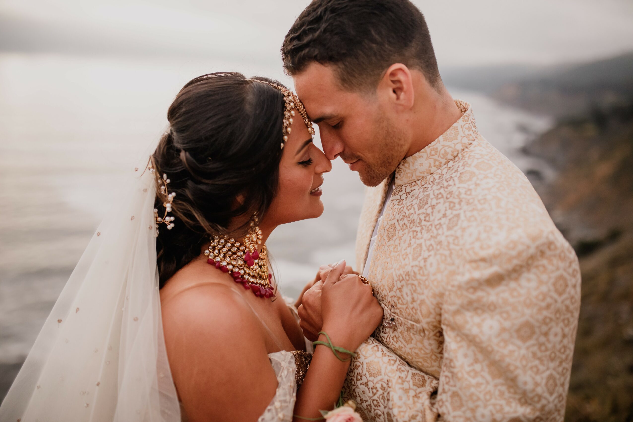 Bride and groom face each other closely outdoors, holding hands, with a blurred natural background.