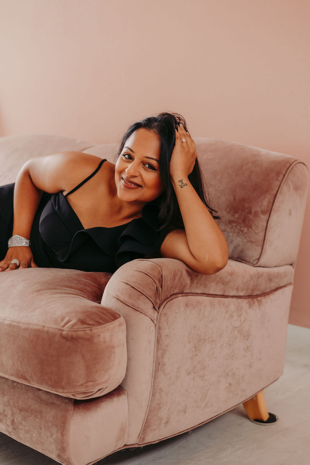 Woman lying on a pink velvet sofa, smiling, resting her head on her hand, wearing a black top, in a relaxed pose.