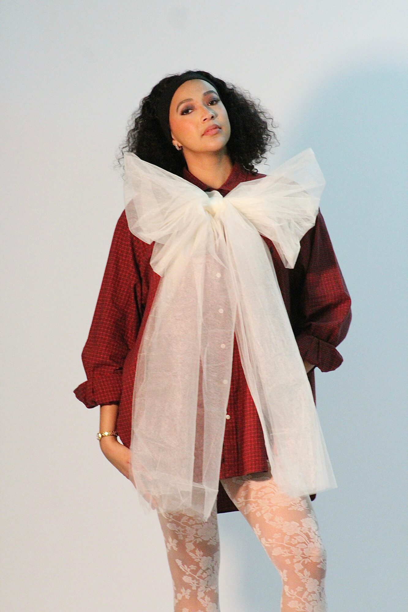 Woman with curly hair wearing a red dress and large white bow, standing against a plain background.