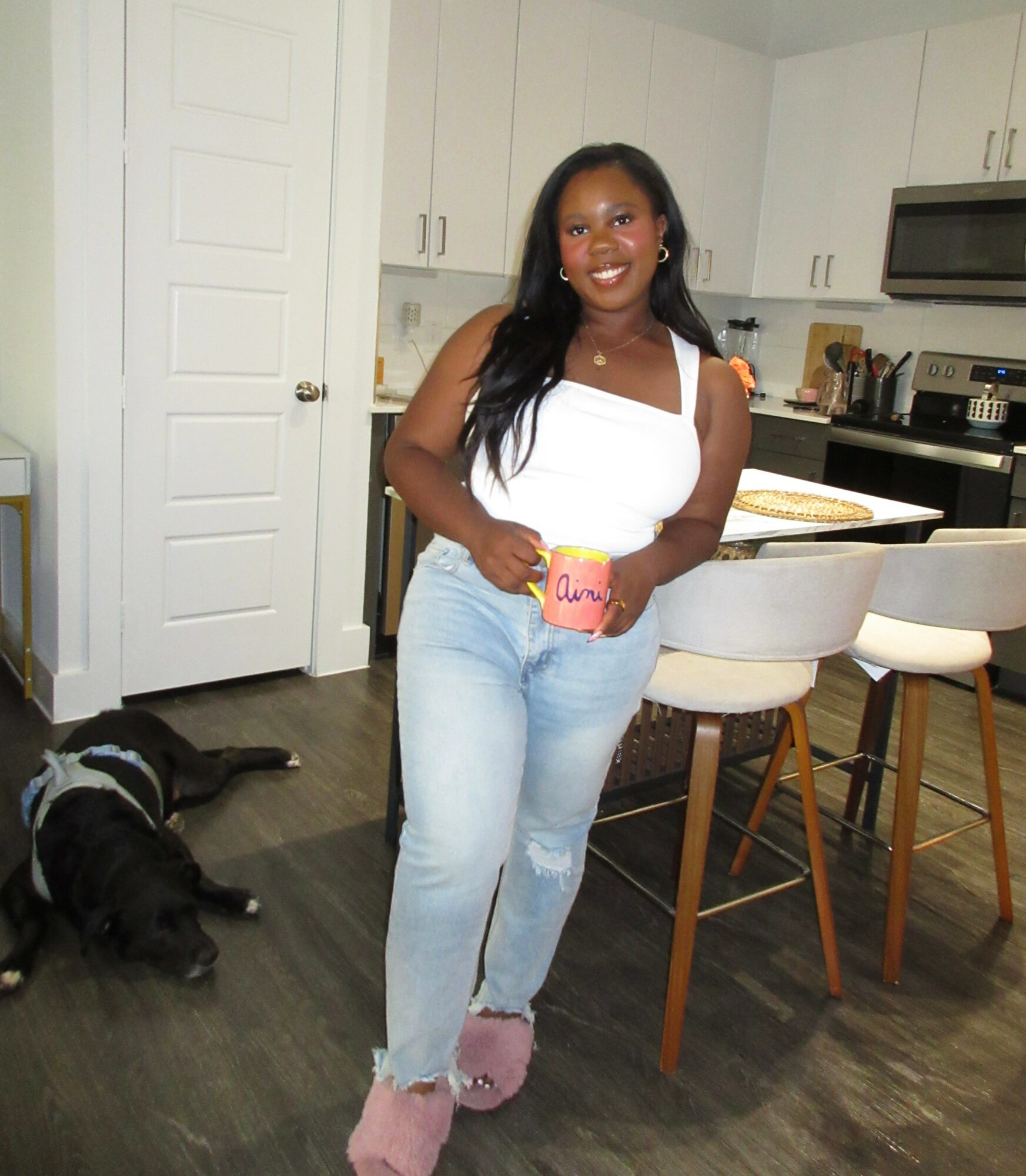 Woman standing in kitchen holding a pink cup, smiling, with a black and white dog lying on the floor nearby.