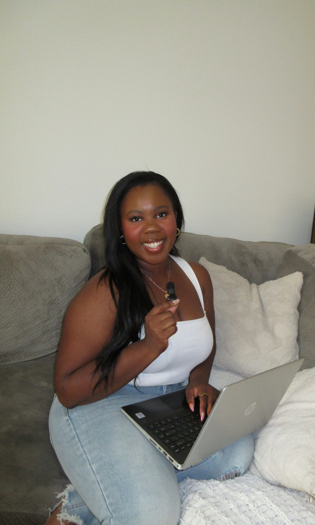 Woman sitting on sofa with laptop, smiling, holding a pen, white top, dark hair, light-colored wall background.