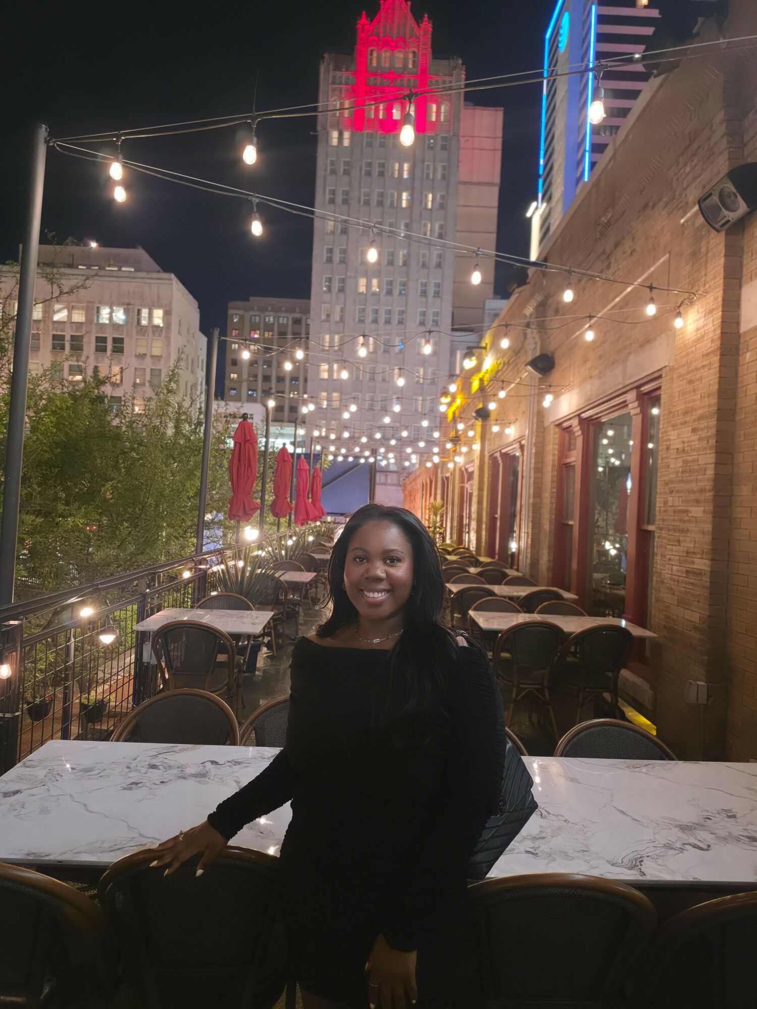 Woman standing in a restaurant with outdoor seating at night, city buildings in the background, illuminated by string lights.
