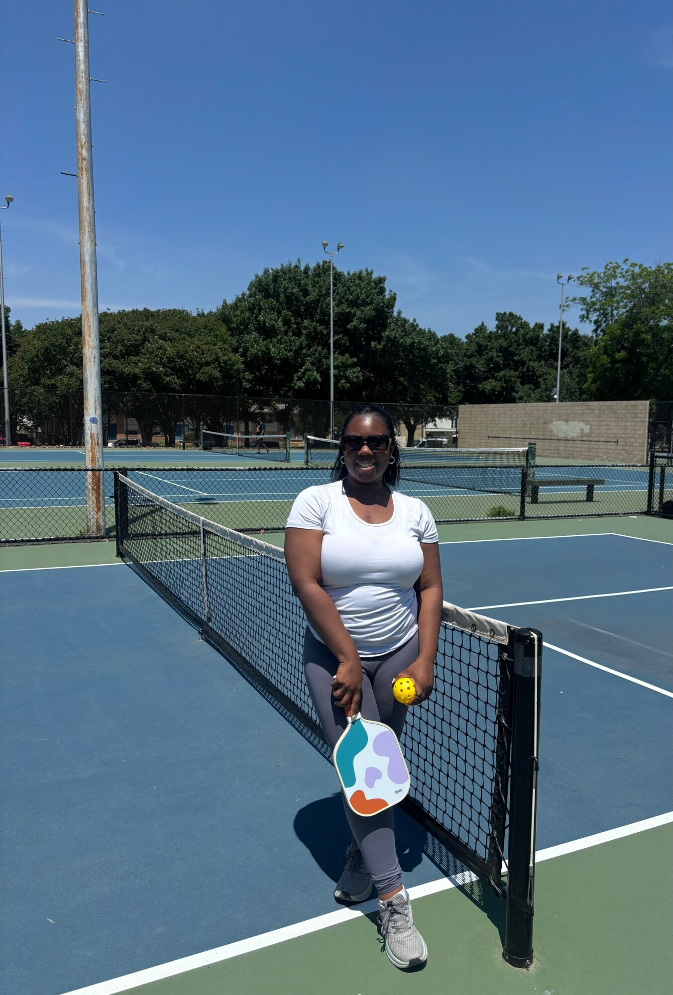 Person standing on a tennis court holding a tennis ball and paddle, with trees and a blue sky in background.