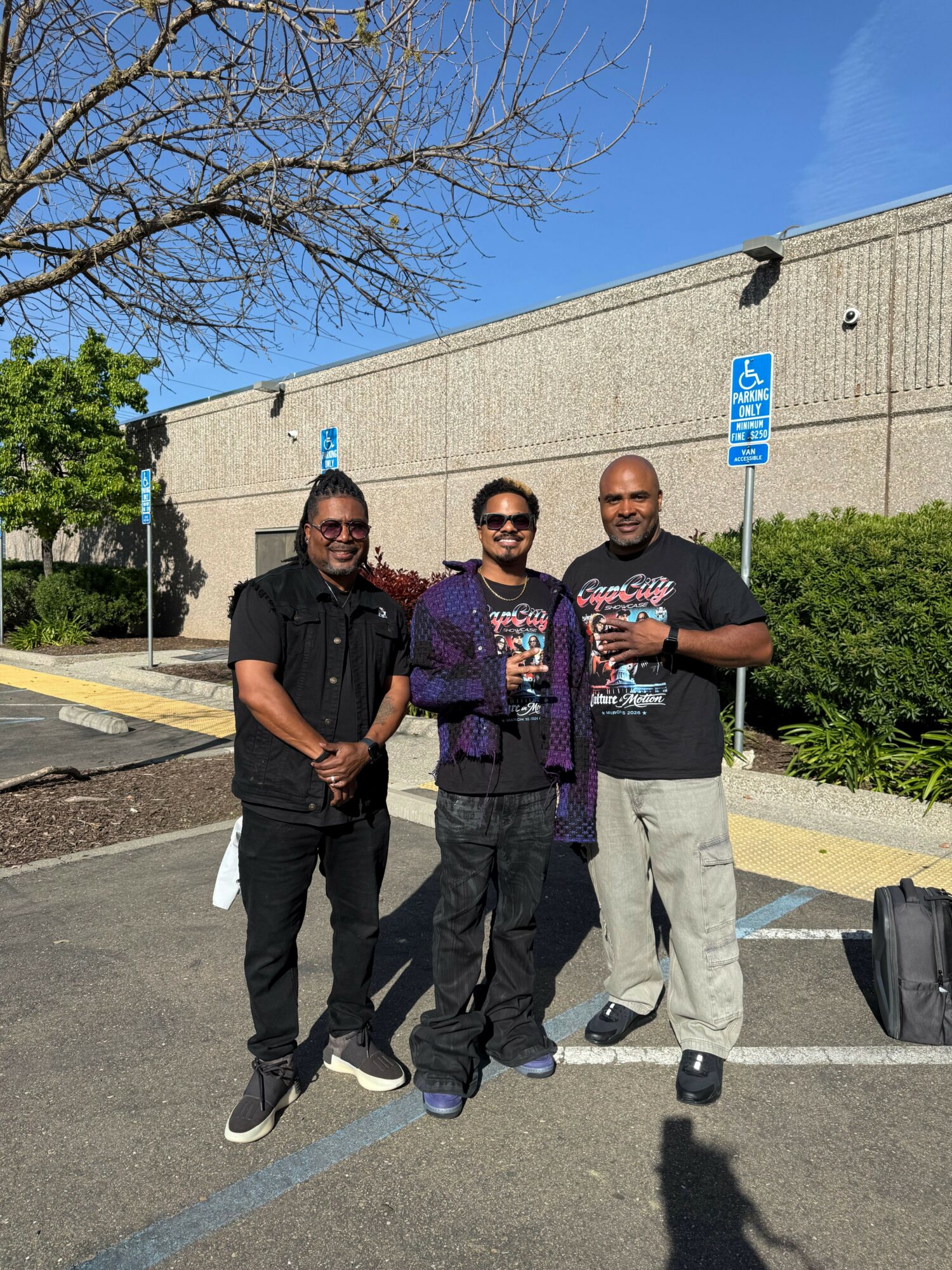 Three people standing outdoors in a parking lot with a building and trees in the background.