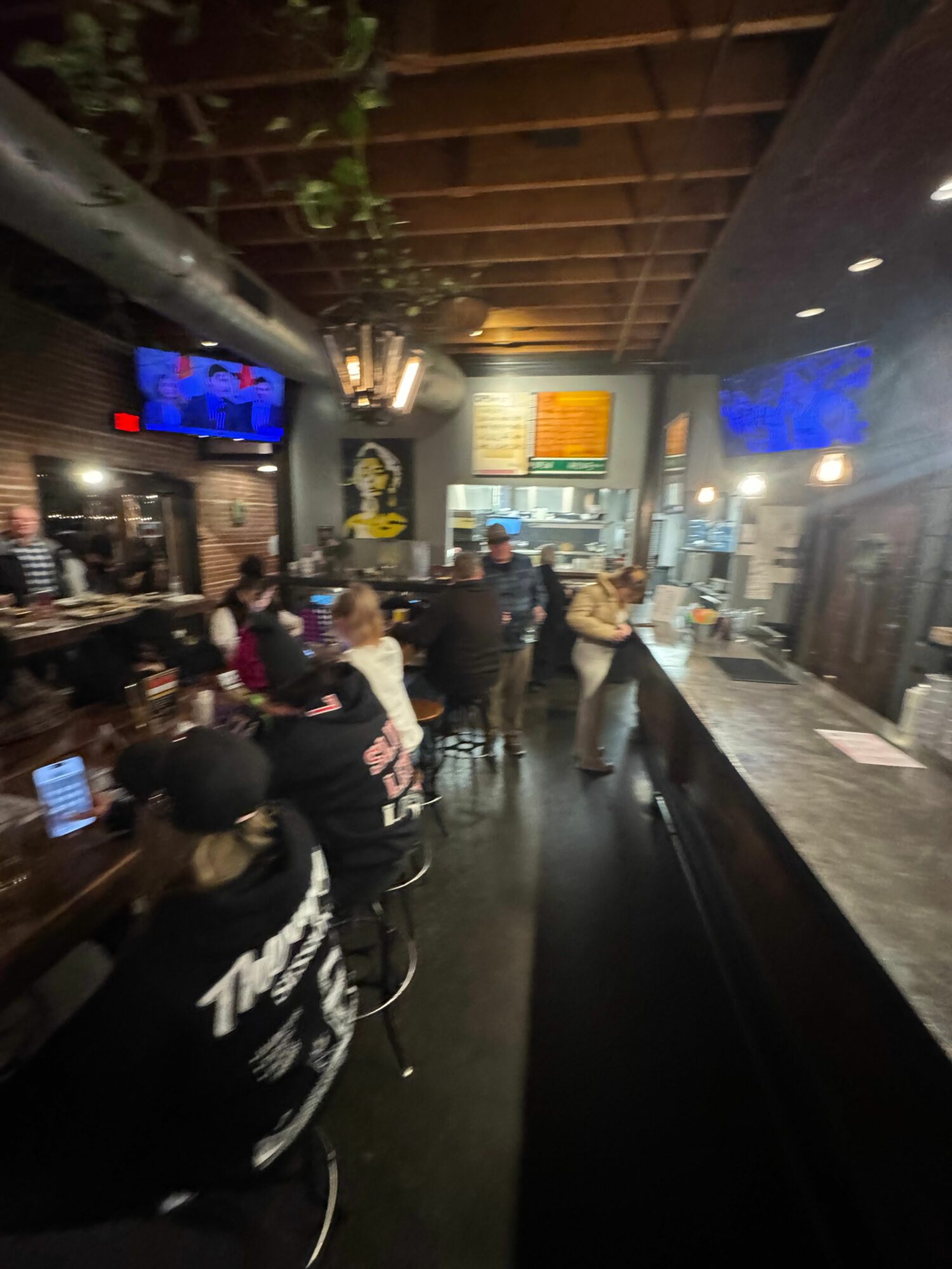 People wait in line at a bar or restaurant with a dark interior, illuminated screens, and a wooden ceiling, during evening.