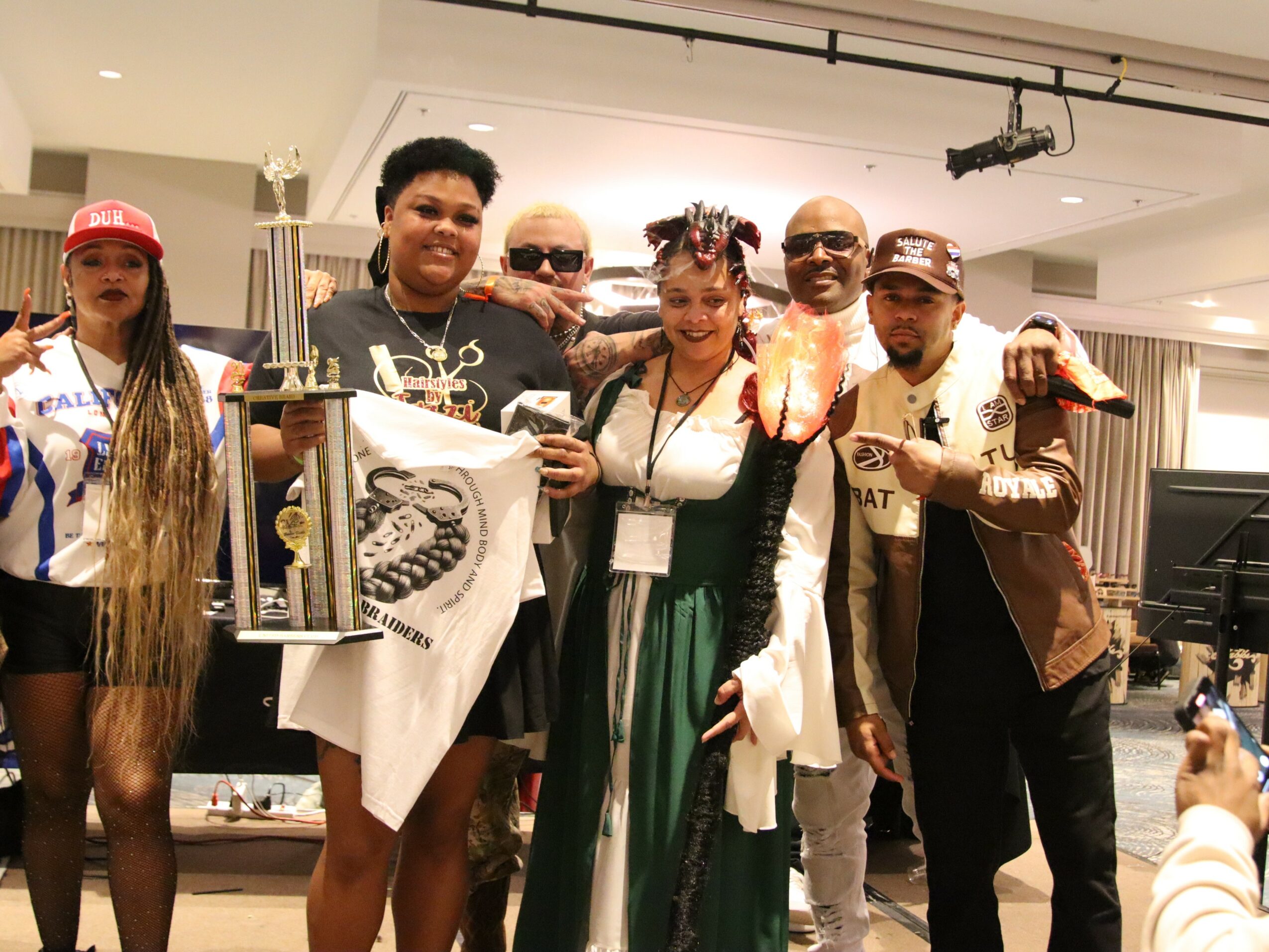 Group of six people smiling indoors, some holding trophies and a flag, with a woman in the center wearing a green dress.