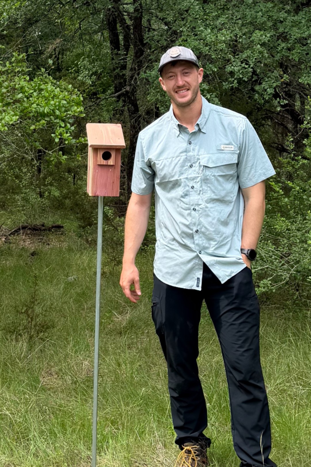 Young man standing outdoors next to a birdhouse on a pole, smiling, surrounded by trees and grass.