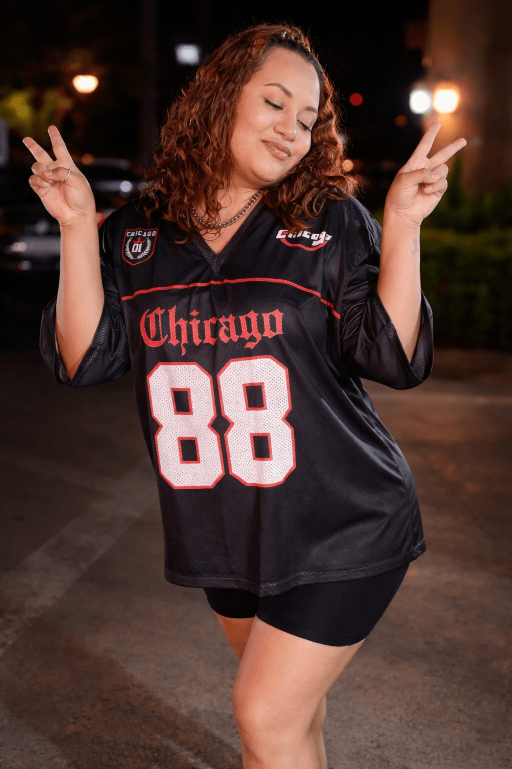 Young woman with curly hair making peace signs with both hands, wearing a black sports jersey with 'Chicago 88' on it, outdoors at night.