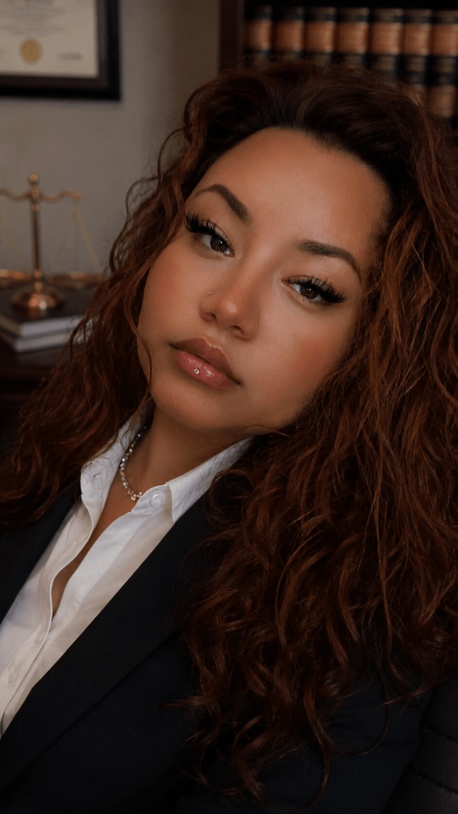 Close-up of a woman with curly hair, wearing a white shirt and dark blazer, in an indoor setting.