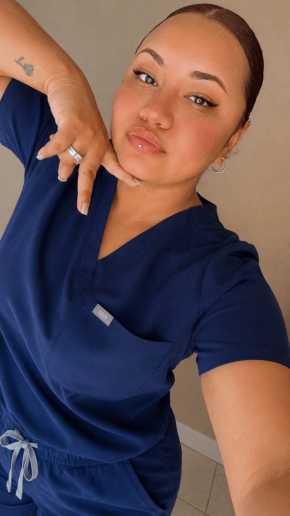 Woman with dark hair in a bun wearing navy scrubs, posing with her hand near her face.