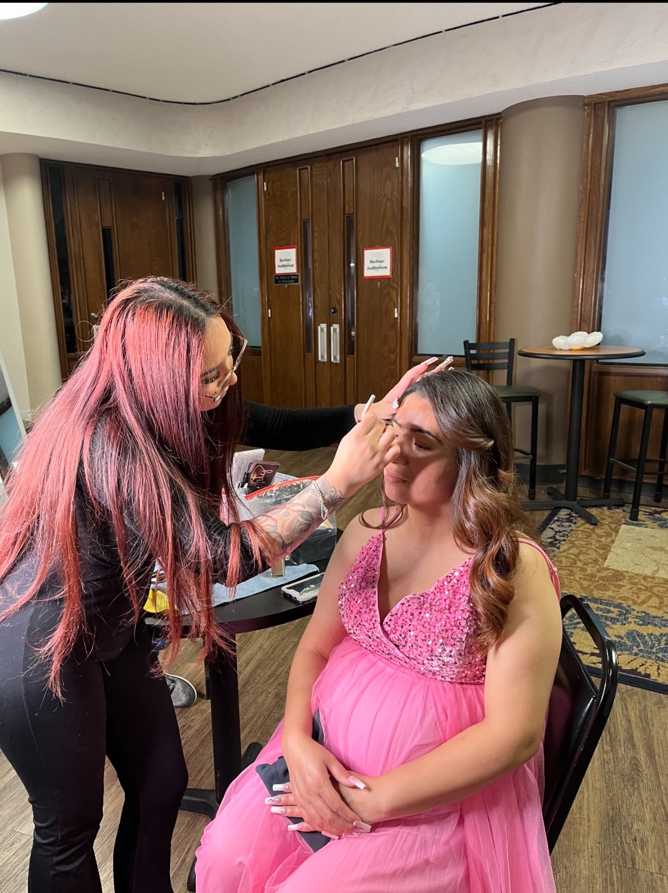 A woman in a pink dress is getting her makeup or face touched up by another woman with long pink hair in a room with wooden doors and chairs.