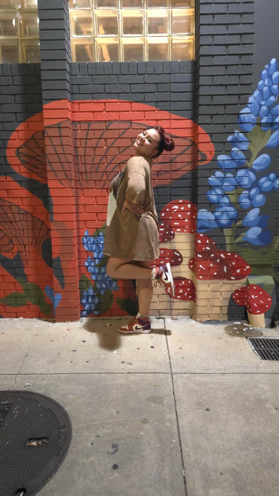 Person standing in front of a colorful mural with flowers and a bench, wearing a beige dress and sunglasses.