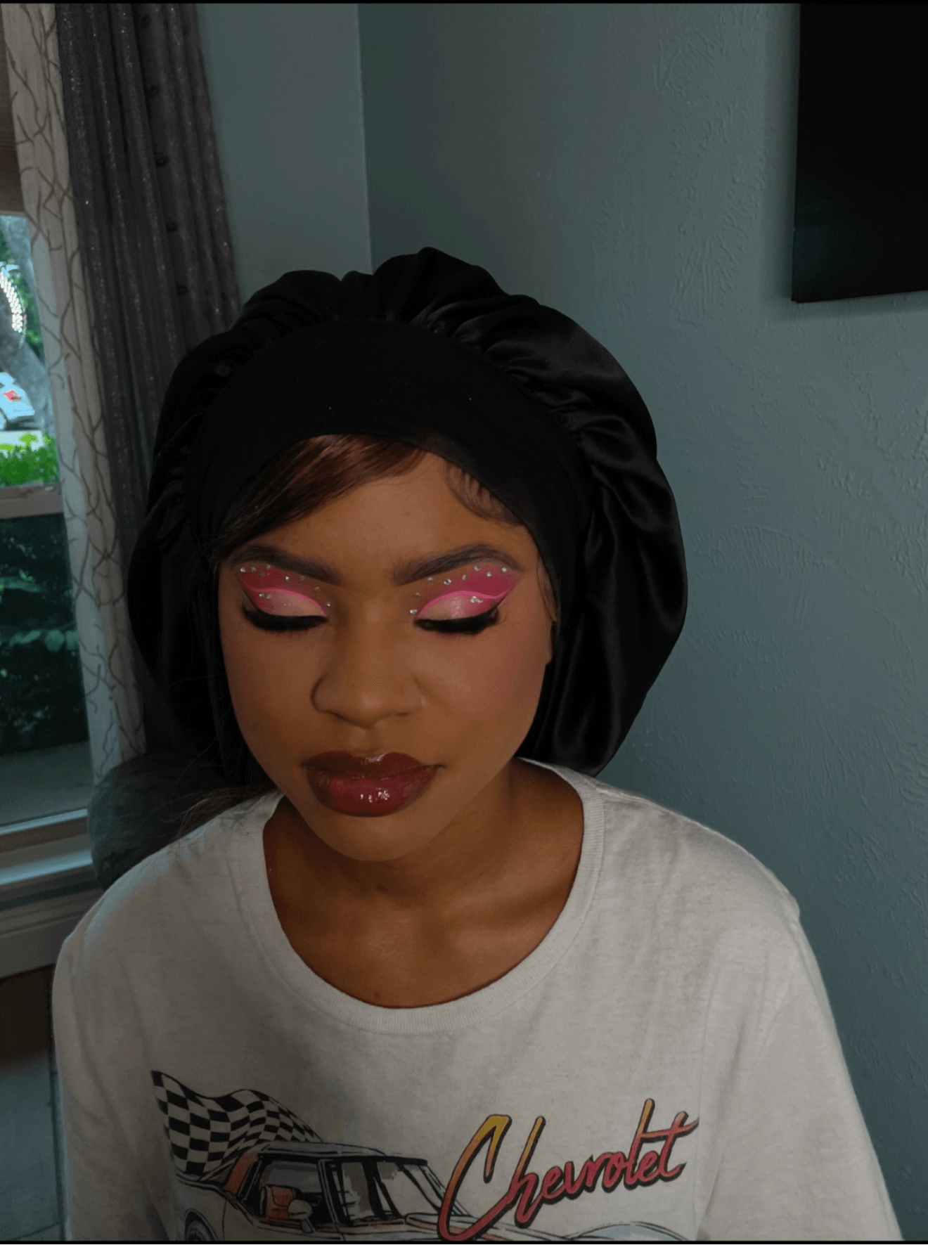 Young woman with pink eyeshadow, dark lipstick, wearing a black headwrap and white shirt, indoors.
