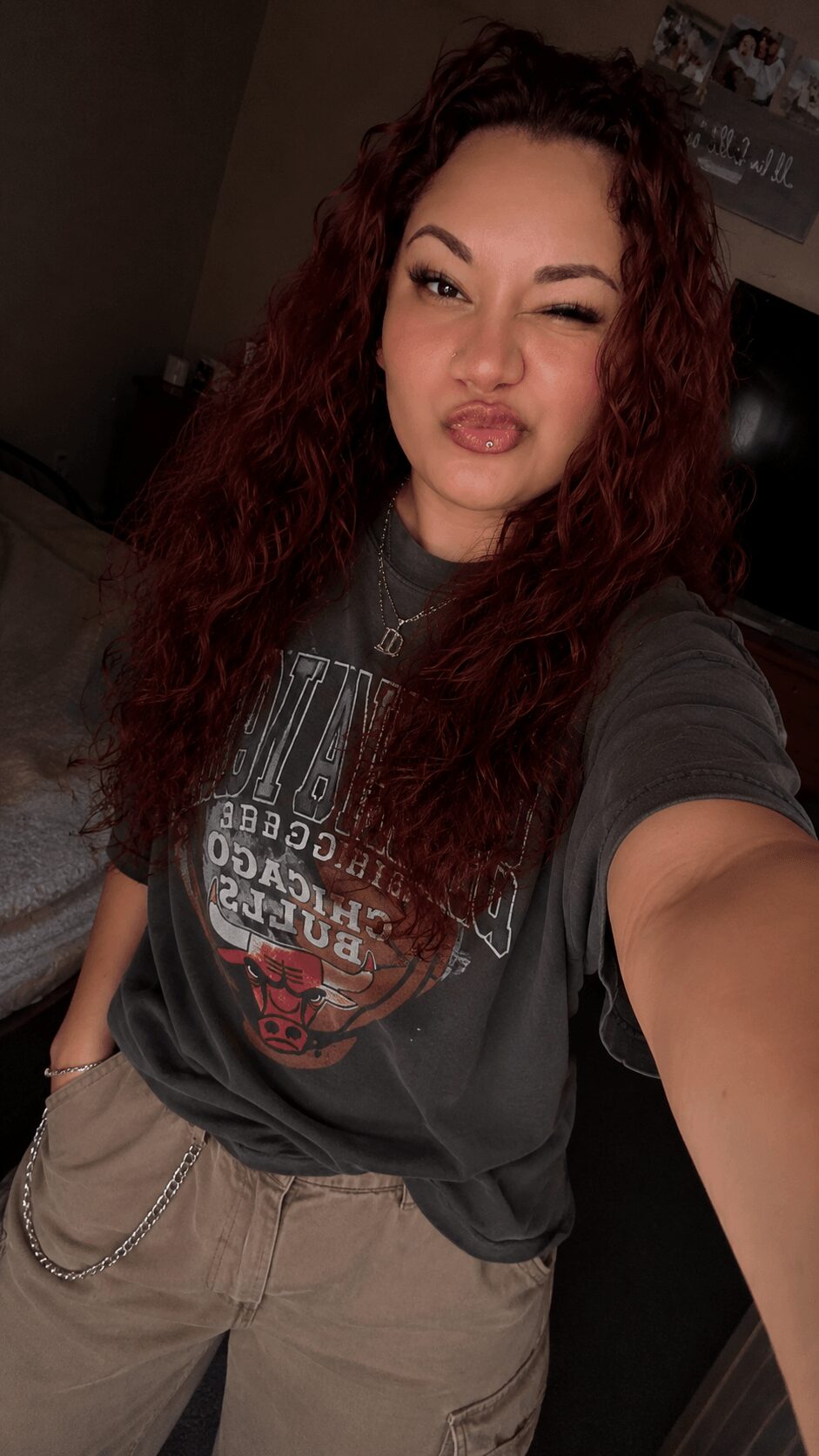 Young woman with curly hair making a playful face, wearing a graphic t-shirt and beige pants, in a dimly lit room.