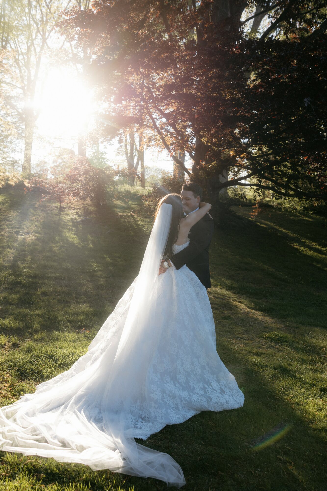 A bride and groom embrace outdoors with sunlight filtering through trees, bride in a white gown with a long train.