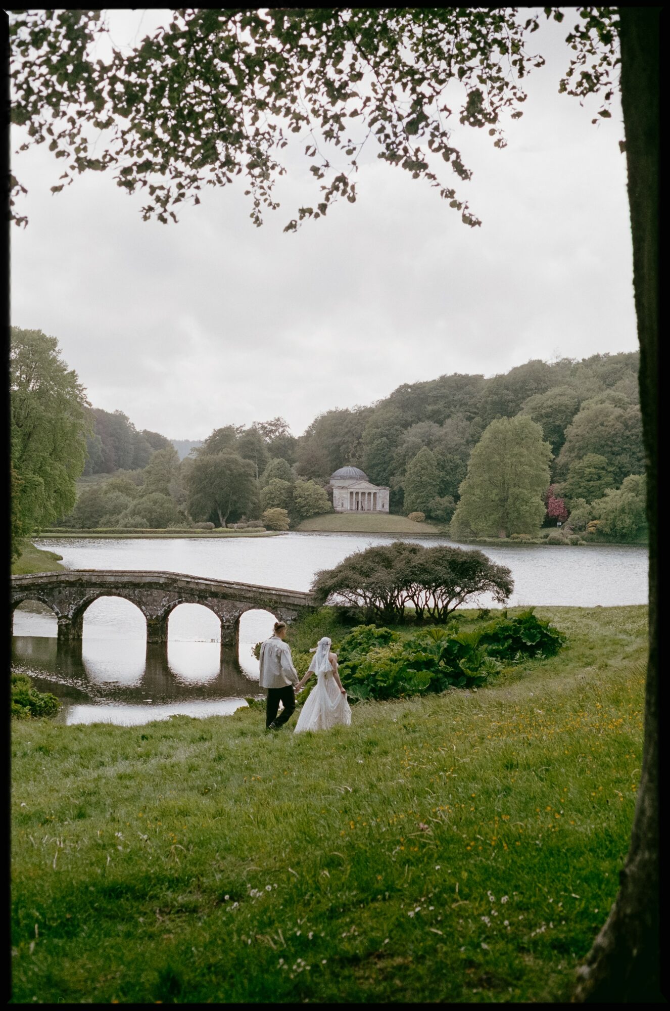 Couple walking near a lake with a bridge, surrounded by trees and a building in the background.