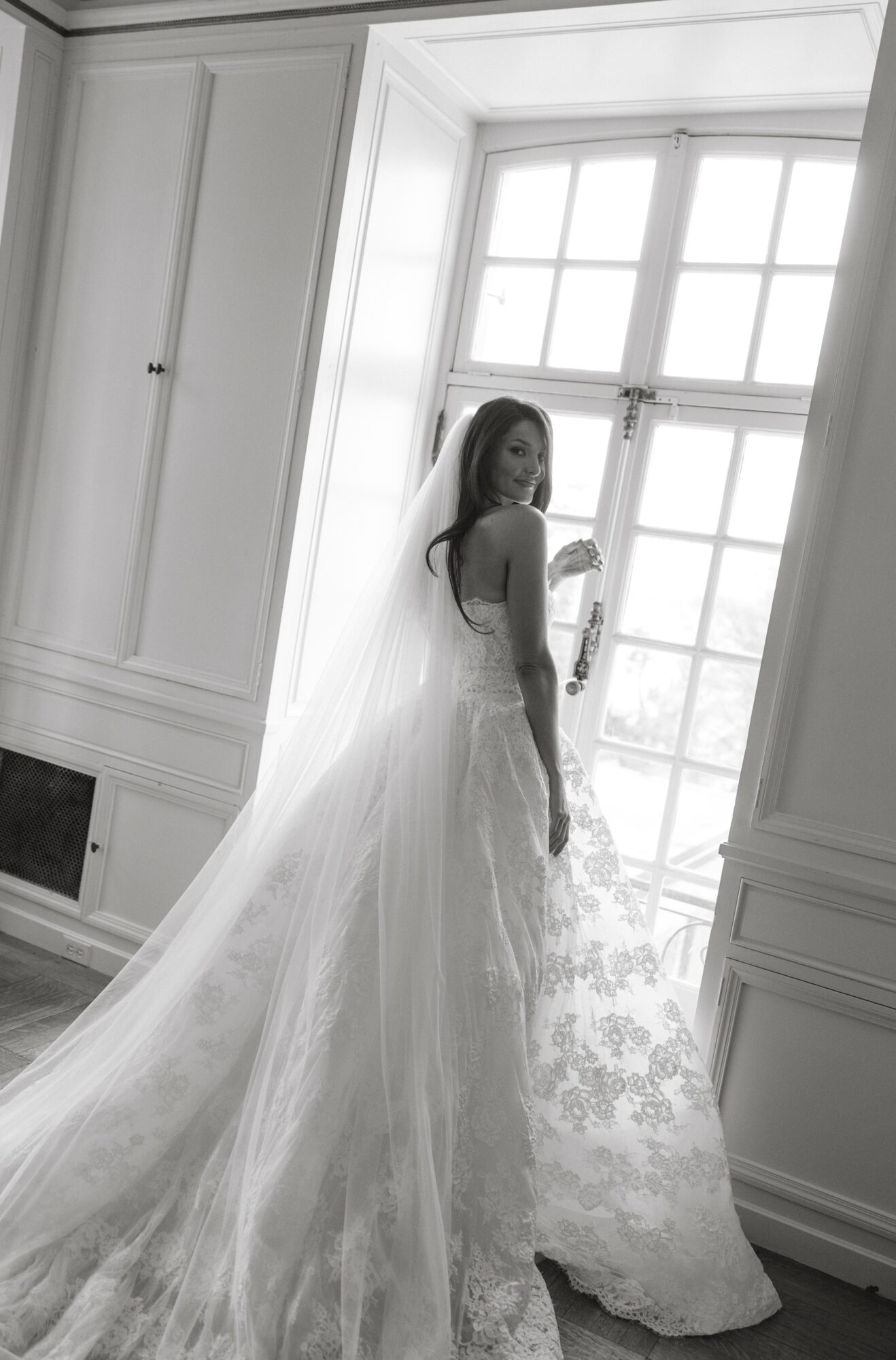 Woman in wedding dress standing by window, looking back, with long train flowing behind her, in a bright room.