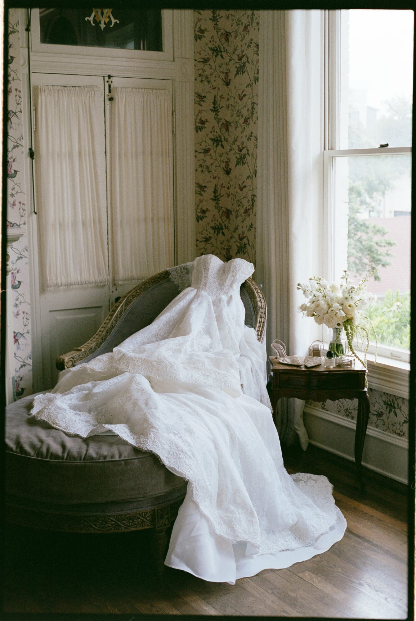 A white wedding dress draped over a vintage sofa in a room with floral wallpaper and large windows, sunlight streaming in.