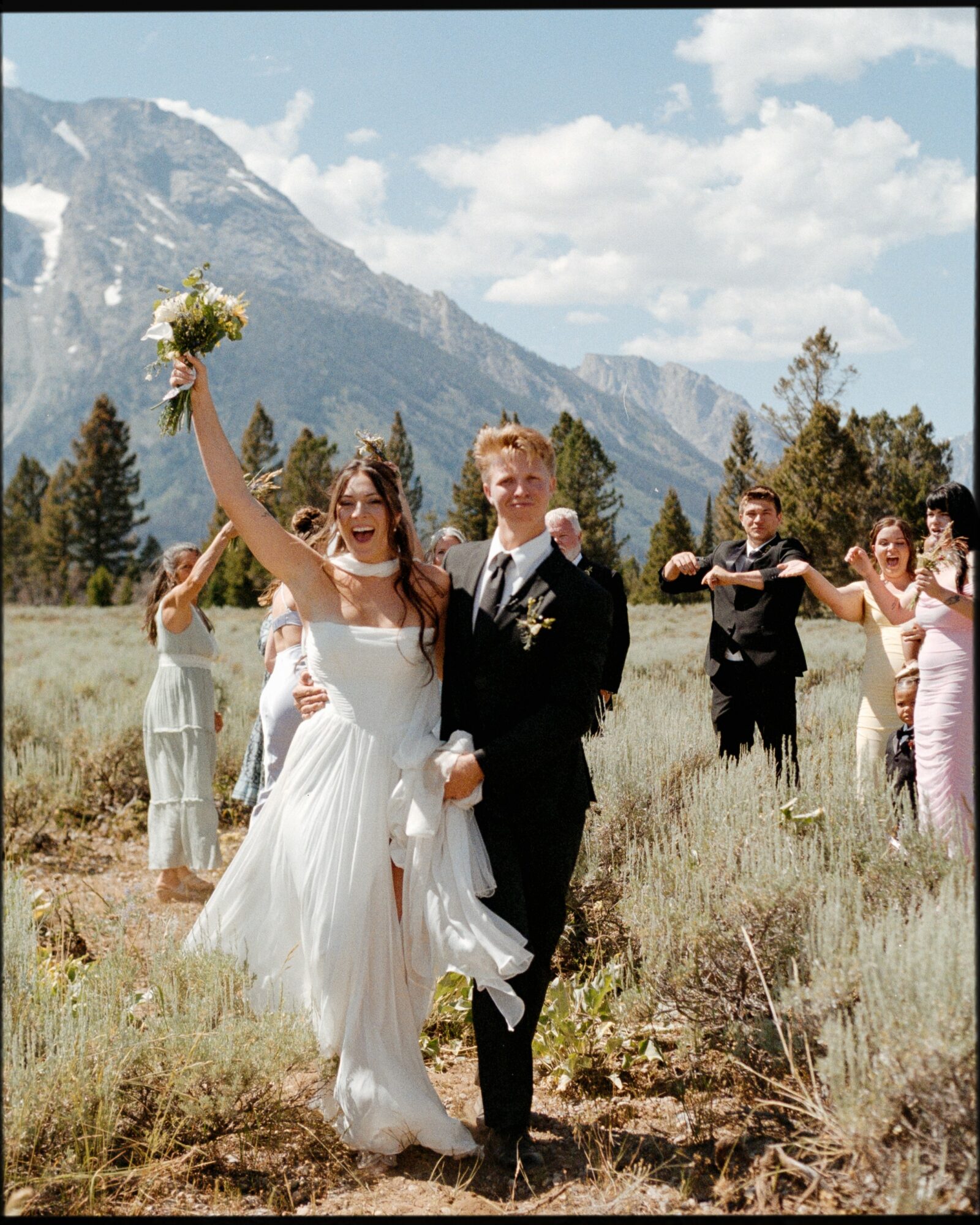 Bride and groom walking outdoors with mountains and trees, bride holding bouquet, guests in background, sunny day.