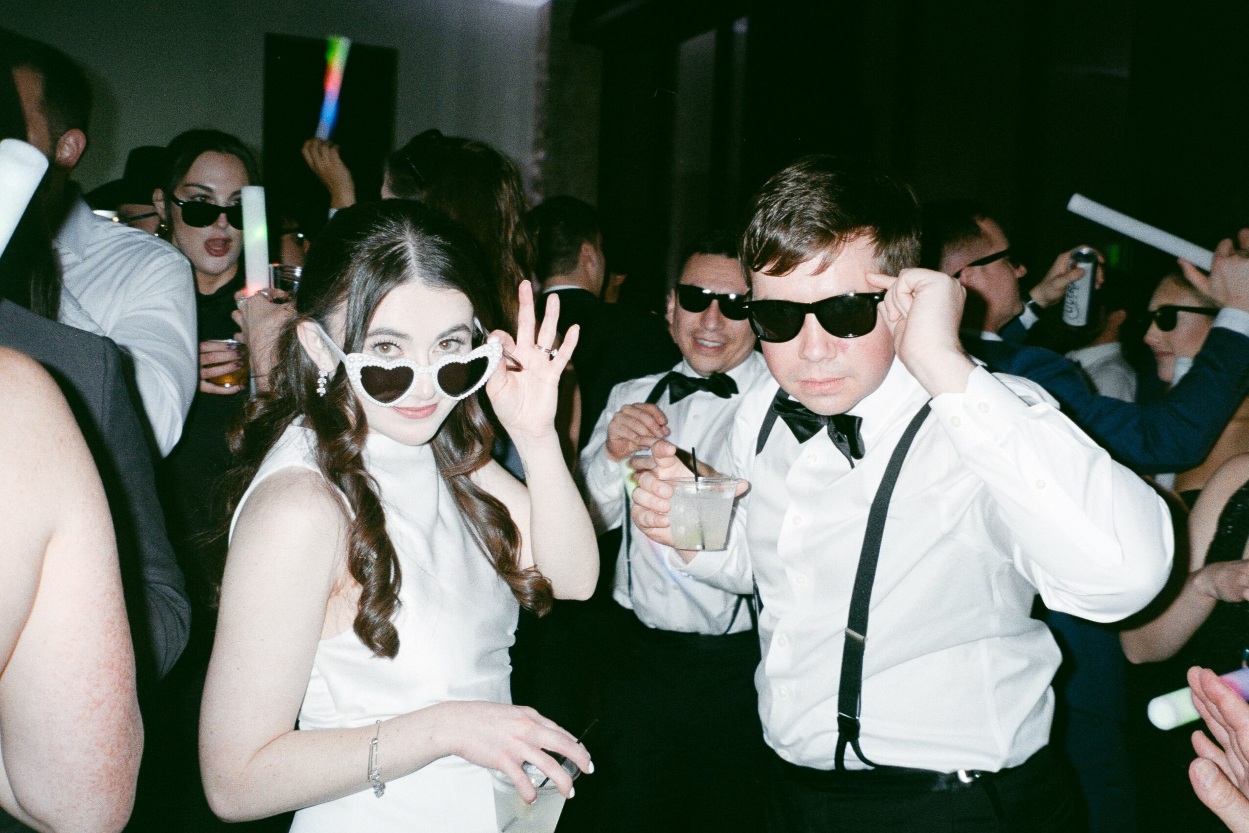 Group of people at a party wearing sunglasses, some waving, with a woman in the foreground and a man adjusting his sunglasses