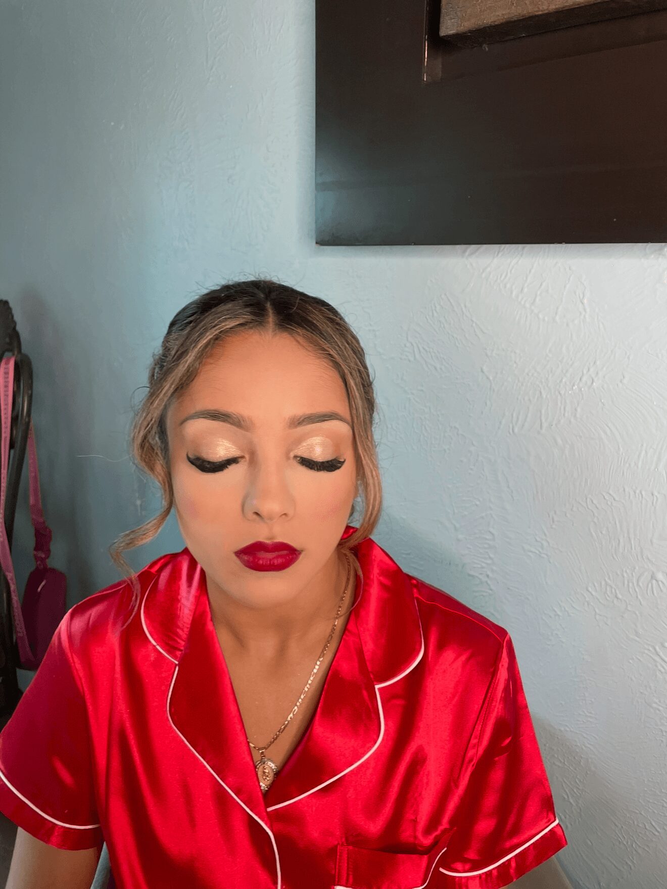 Woman with makeup and red satin robe, closed eyes, in a room with light-colored wall and dark frame, listening to earphones.