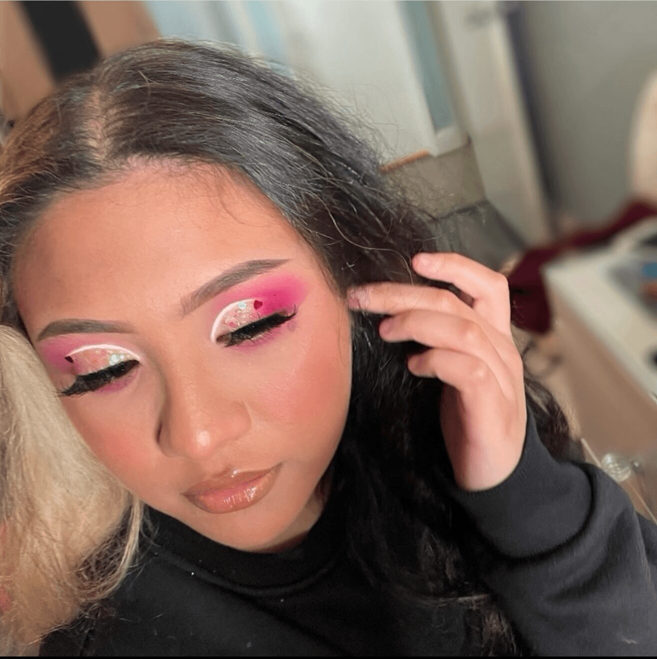 Close-up of a woman with colorful eye makeup, pink and white eyeshadow, and glossy lips, touching her hair.