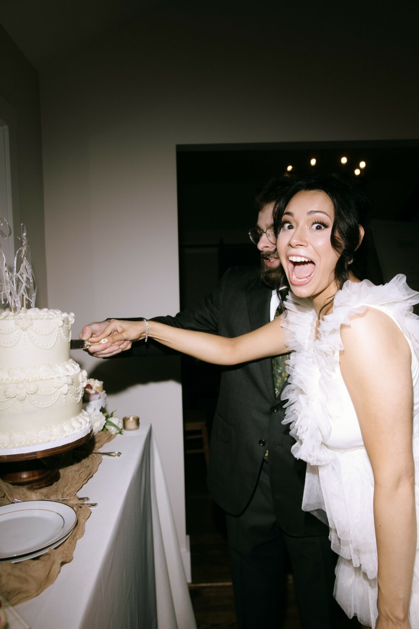 Woman in wedding dress smiling and reaching for a cake, with a person in dark clothing behind her, in a room.