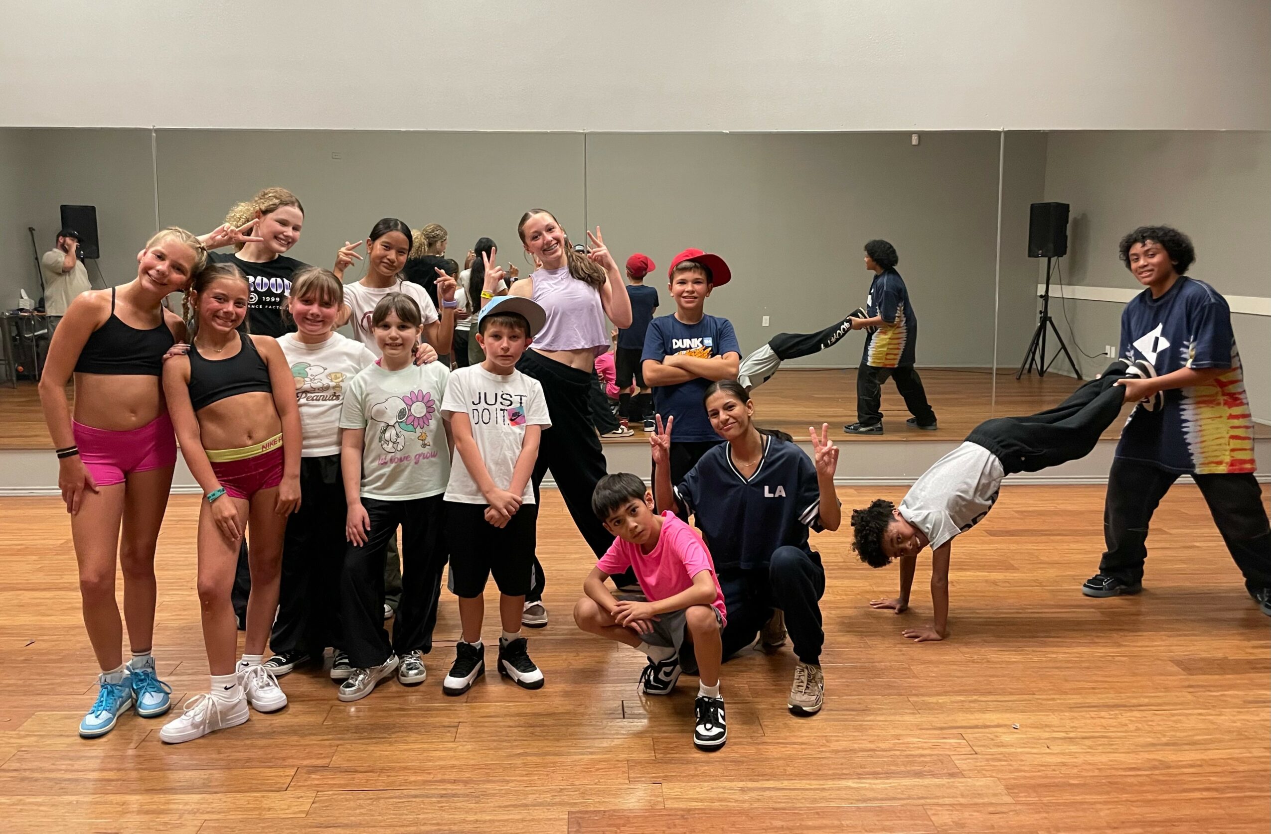 Group of children and teenagers in a dance studio, some posing and others performing breakdancing moves, with a mirror and speakers in the background.