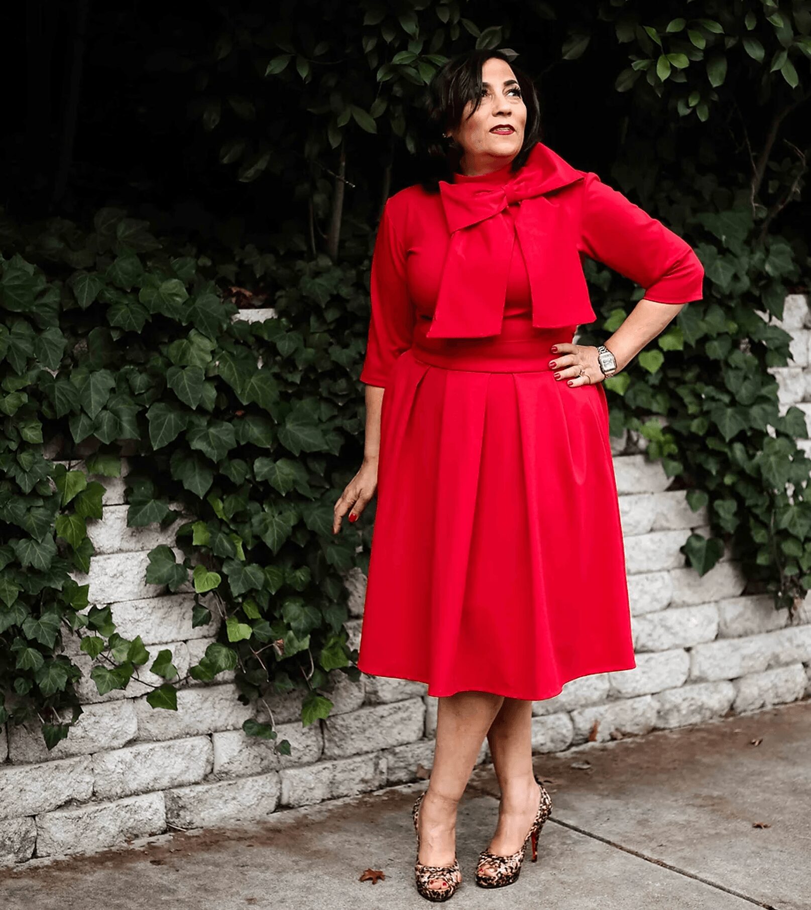 Woman in red dress and cape standing outdoors near a white brick wall with greenery, looking to the side.