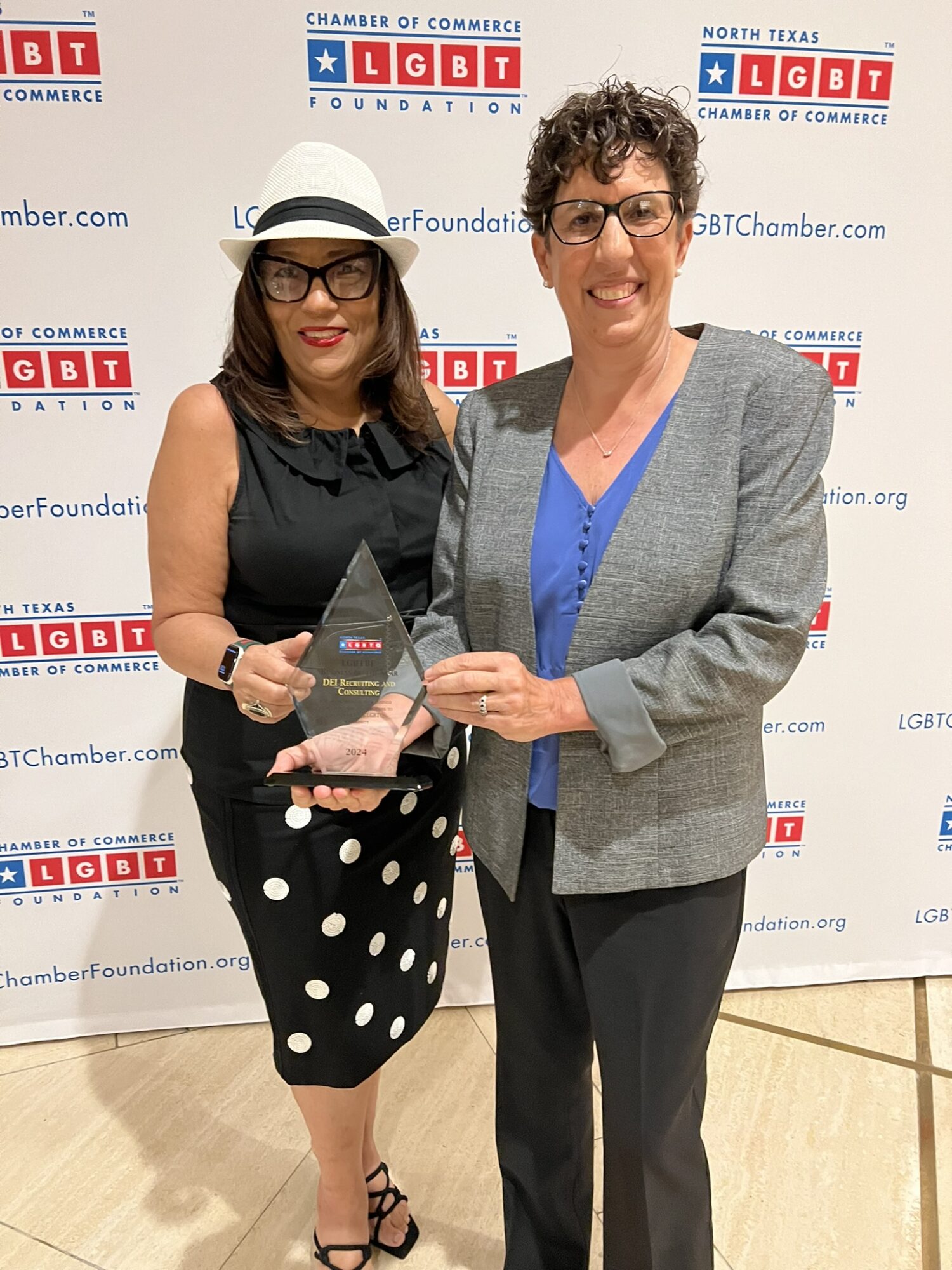 Two women standing together, holding an award, smiling, in front of a backdrop with logos and text, one wearing a hat and glasses.