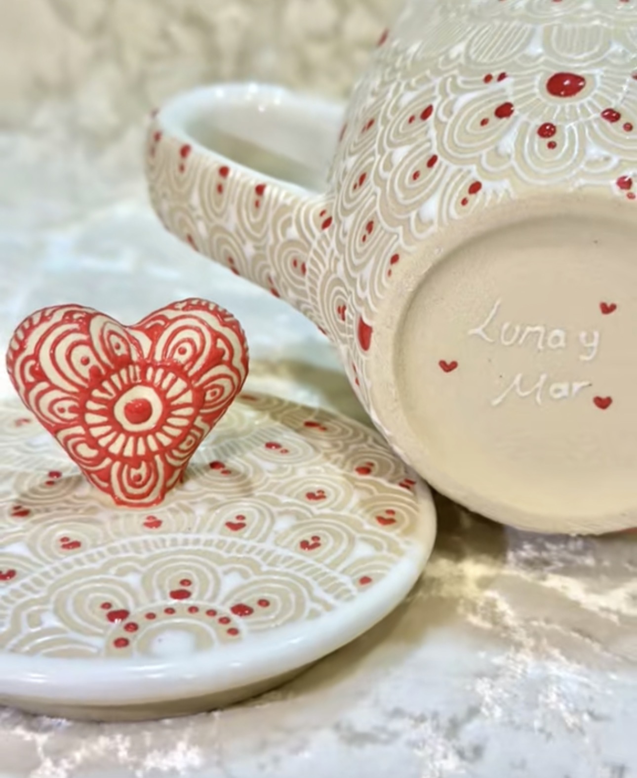 Decorative ceramic mug and plate with red and white swirl patterns, heart-shaped object, and handwritten message, on a marble surface.