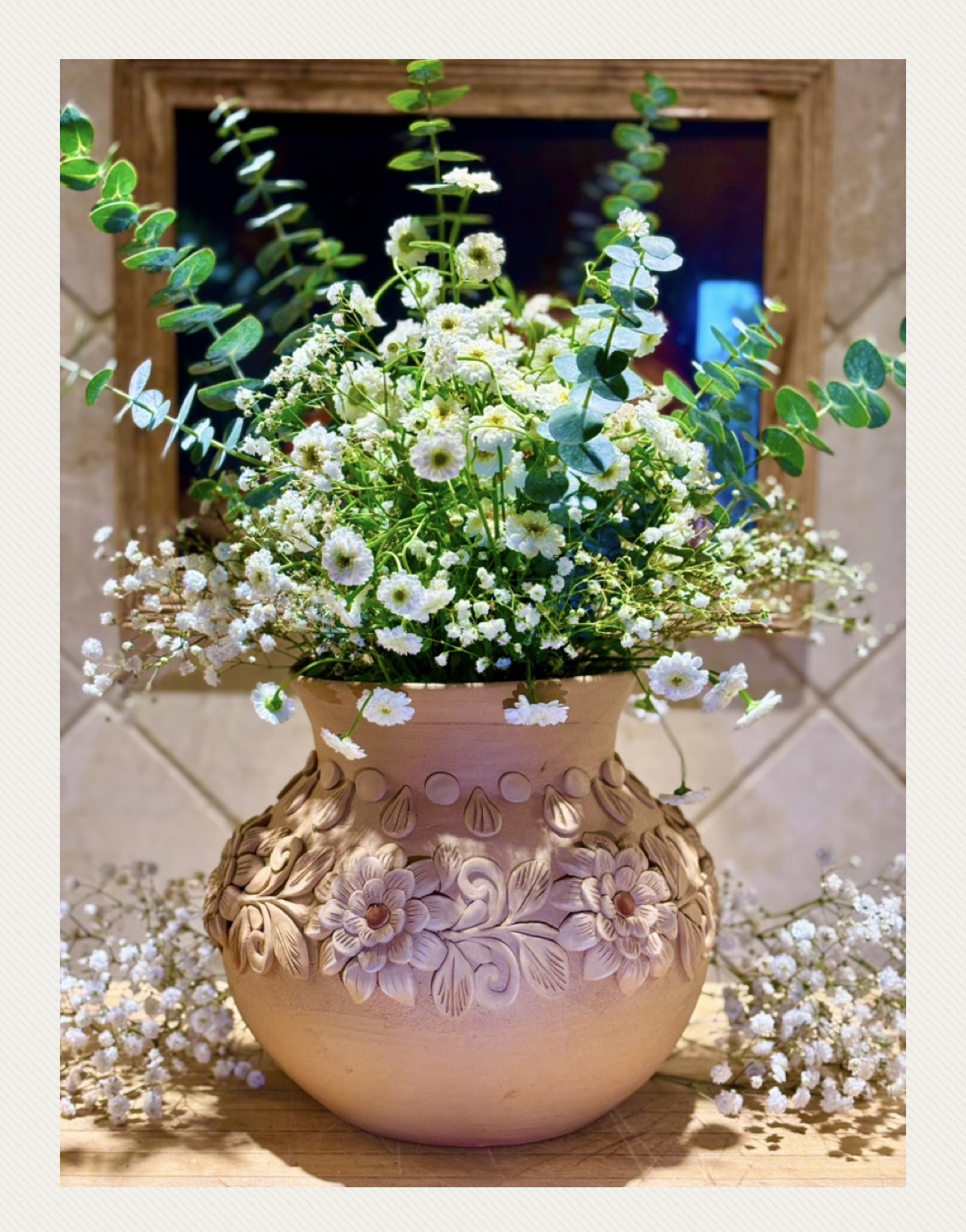 A floral arrangement with white daisies and greenery in a decorative pink vase, placed on a surface with additional flowers around.