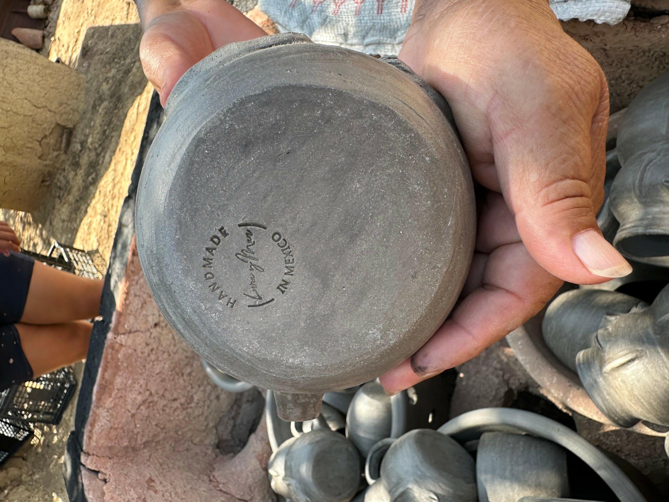 Person holding a gray ceramic bowl with maker's mark on bottom, outdoors with other pottery items nearby.