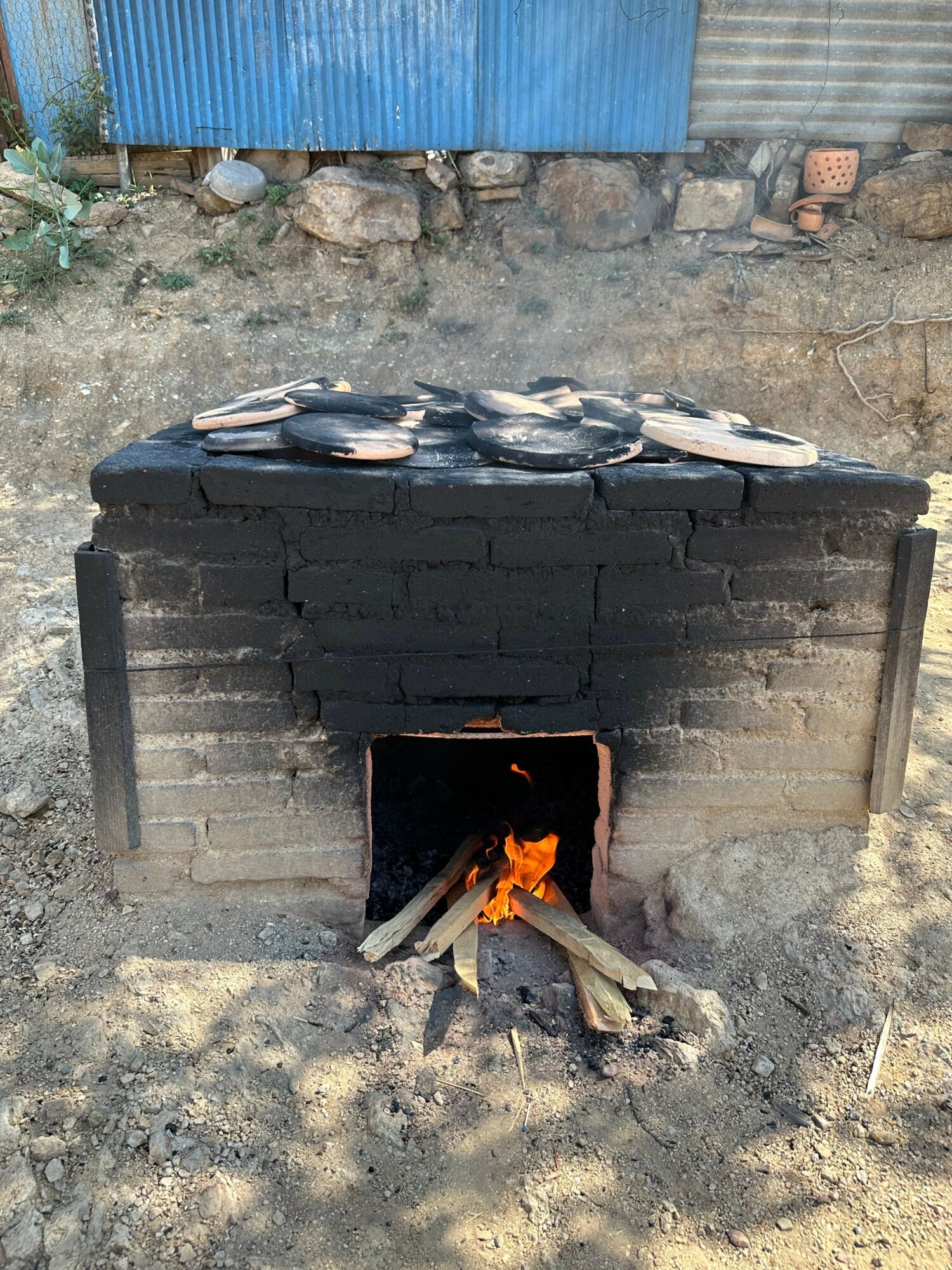 A brick oven with a fire inside, outdoors on dirt ground, with a blue wall and rocks nearby.
