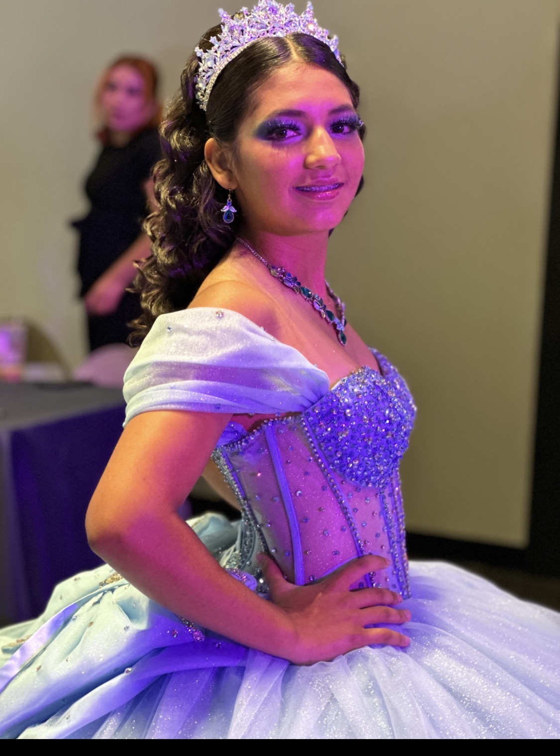 Young woman in a princess dress with a tiara, jewelry, and makeup, posing with hand on hip at event.