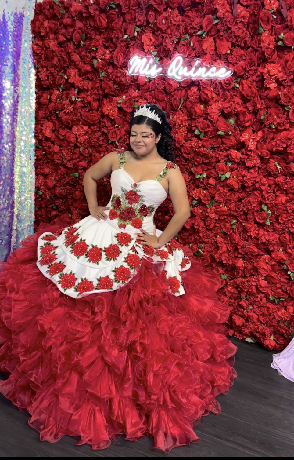 Young woman in a red and white floral dress with a full skirt, standing in front of a red flower wall, smiling.