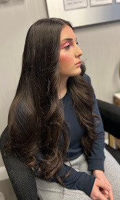Young woman with long, wavy dark hair, wearing a dark top, sitting indoors with a neutral background.