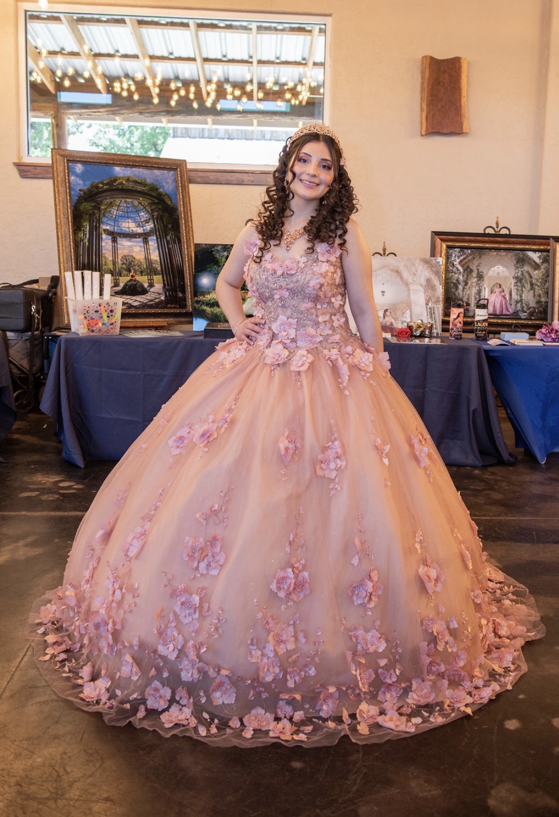 Young woman in a pink floral ball gown with a full skirt, standing indoors with artwork and tables behind her.
