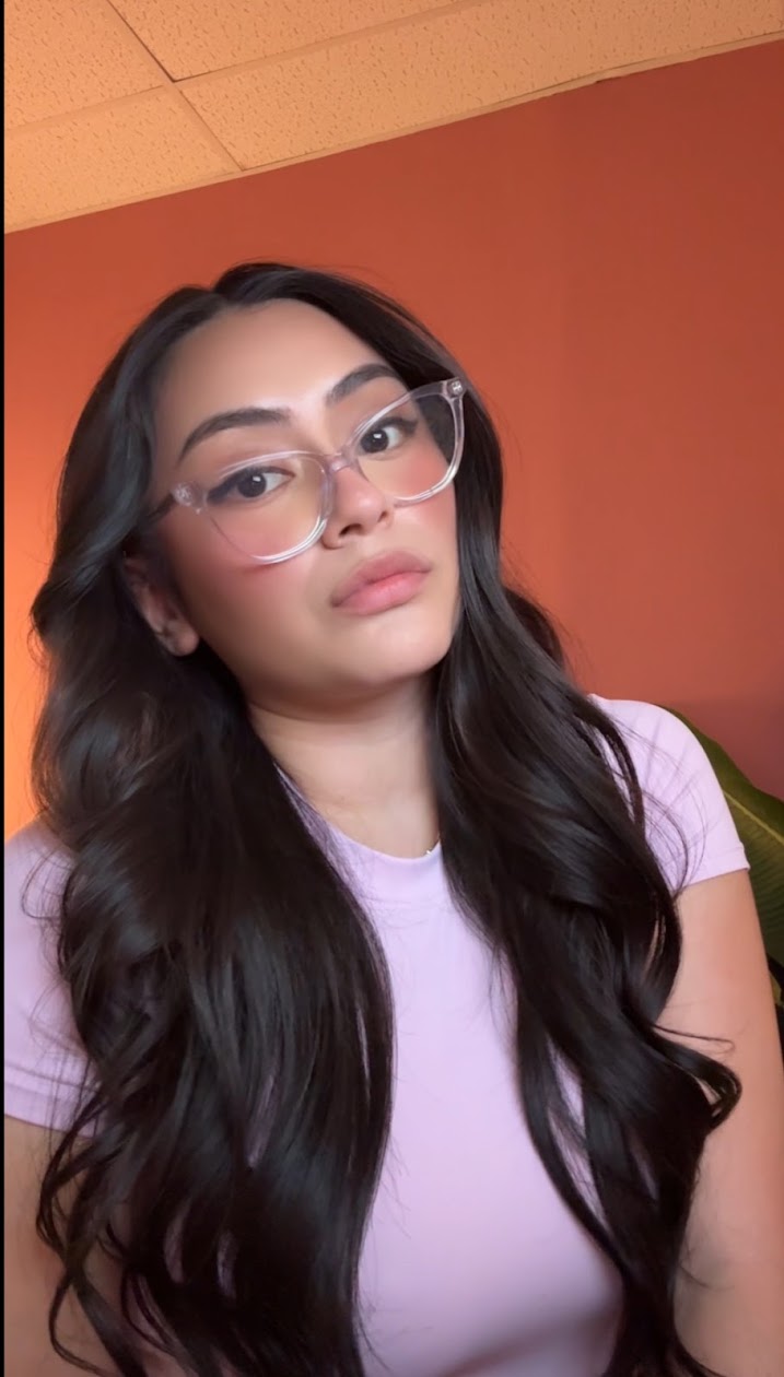 Young woman with long dark hair and glasses, wearing a light purple shirt, against an orange wall.