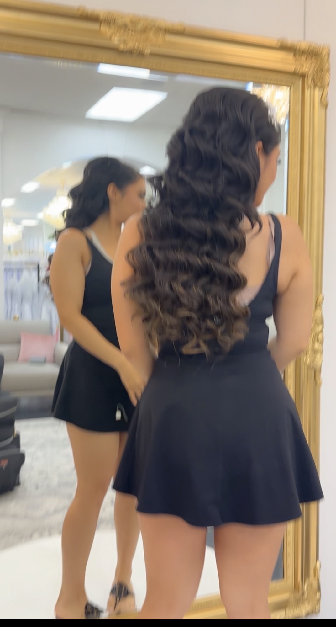 Two women in black dresses standing in front of a large mirror, one facing away and the other reflected, in a room with bright lighting.
