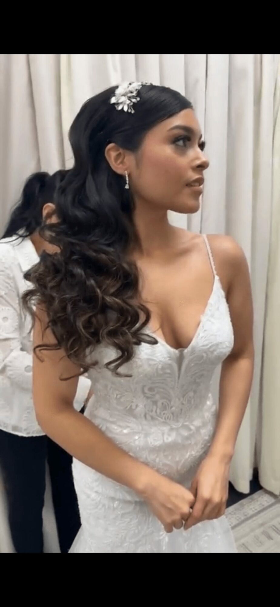 Woman with long, wavy hair wearing a white dress and hair accessory, standing in front of curtains.