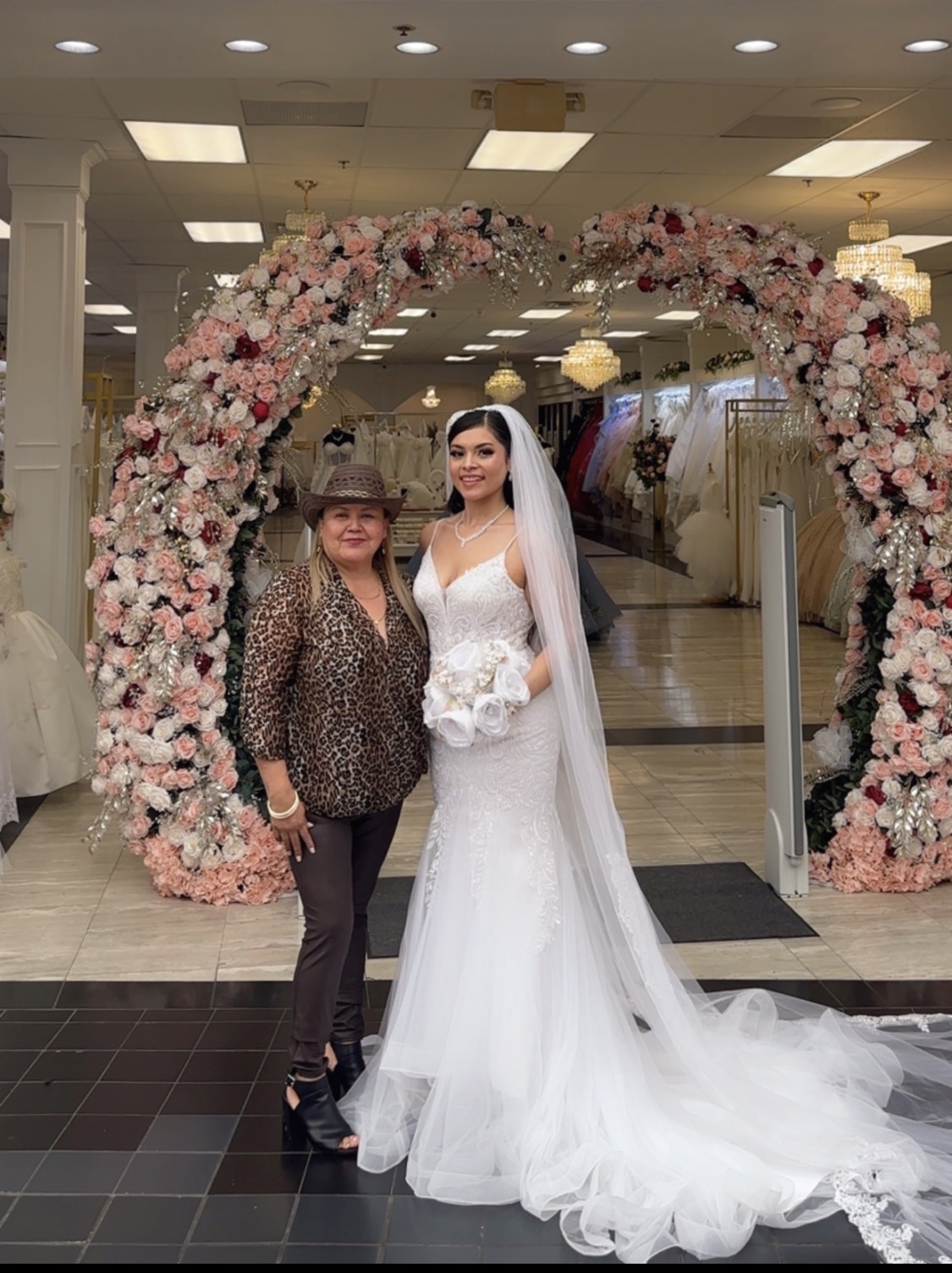 A bride in a white wedding gown and veil stands next to a woman in a leopard print top and hat, in front of a floral arch.