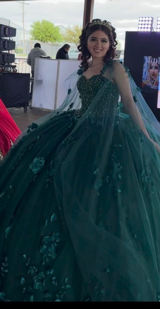Young woman in a green gown smiling at the camera, indoors with people and equipment in the background.