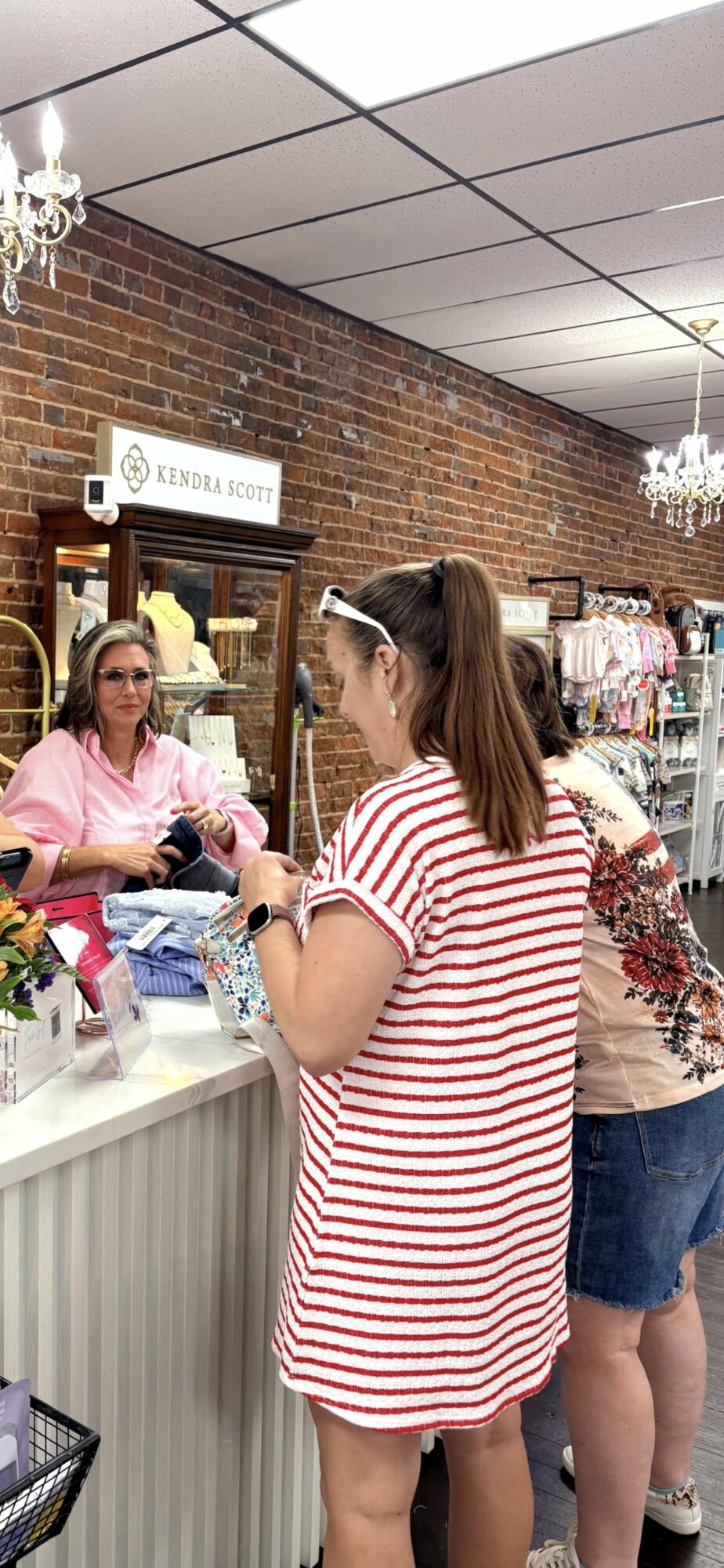 Two women at a store counter, one in a striped dress and the other in a pink shirt, with shelves behind them.