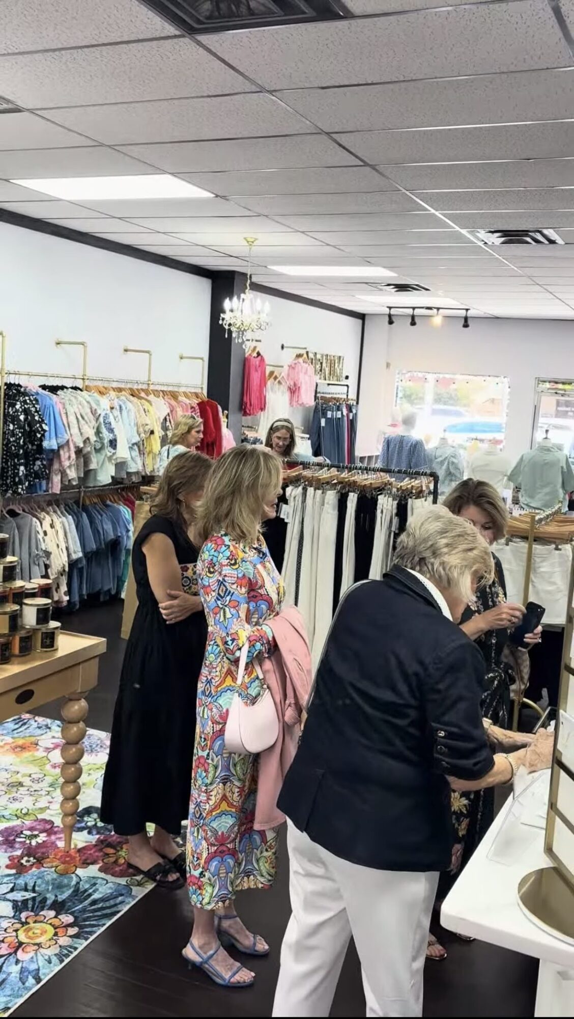 People shopping in a clothing store with racks of colorful clothes and accessories, ceiling lights, and large windows.
